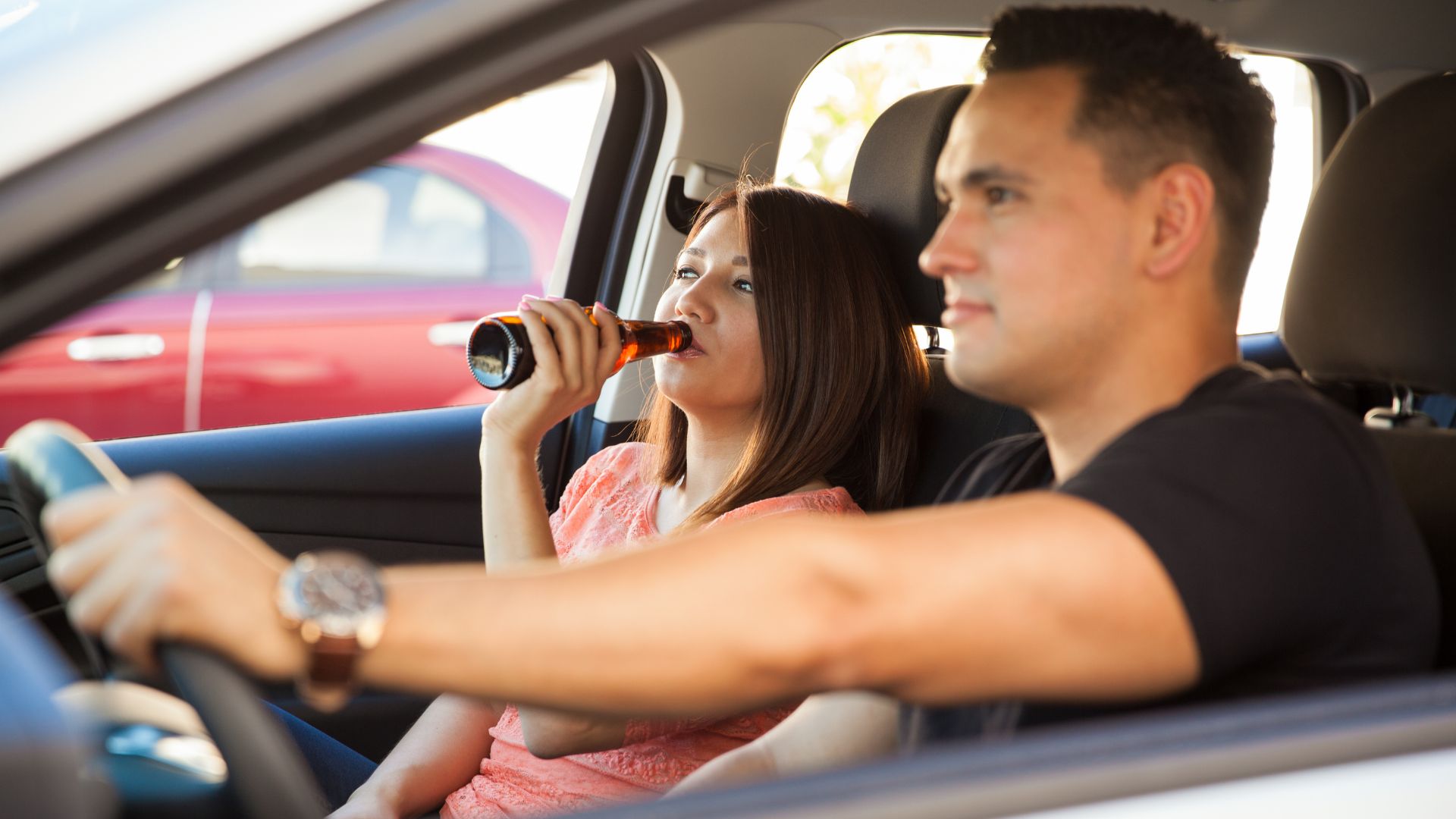 A woman drinking in a car as a passenger while a man is driving her.