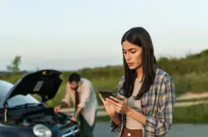 A woman on her phone while her friend is working on a broken car in the background.