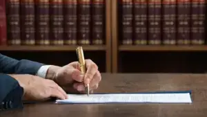 A man signing a paper with a book case of law books in the background.