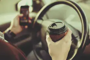 A person behind the wheel with a phone and coffee in each hand.